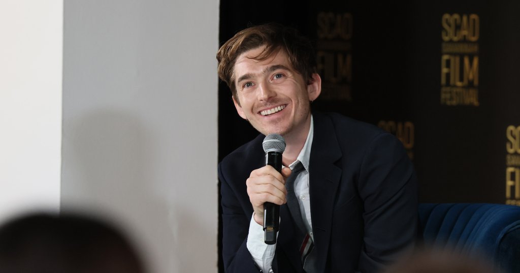 Austin Abrams smiling and holding a microphone while seated onstage at the SCAD Savannah Film Festival.