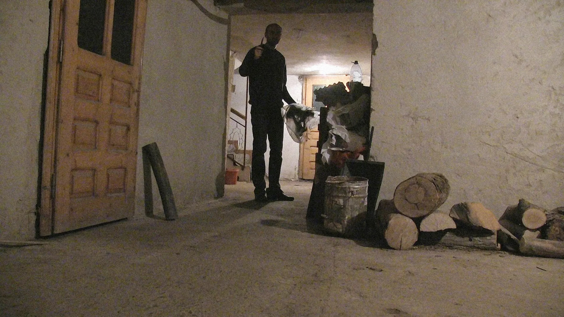 A man in black stands silhouetted in a dimly lit doorway next to stacked firewood and a metal bucket in a run-down room.