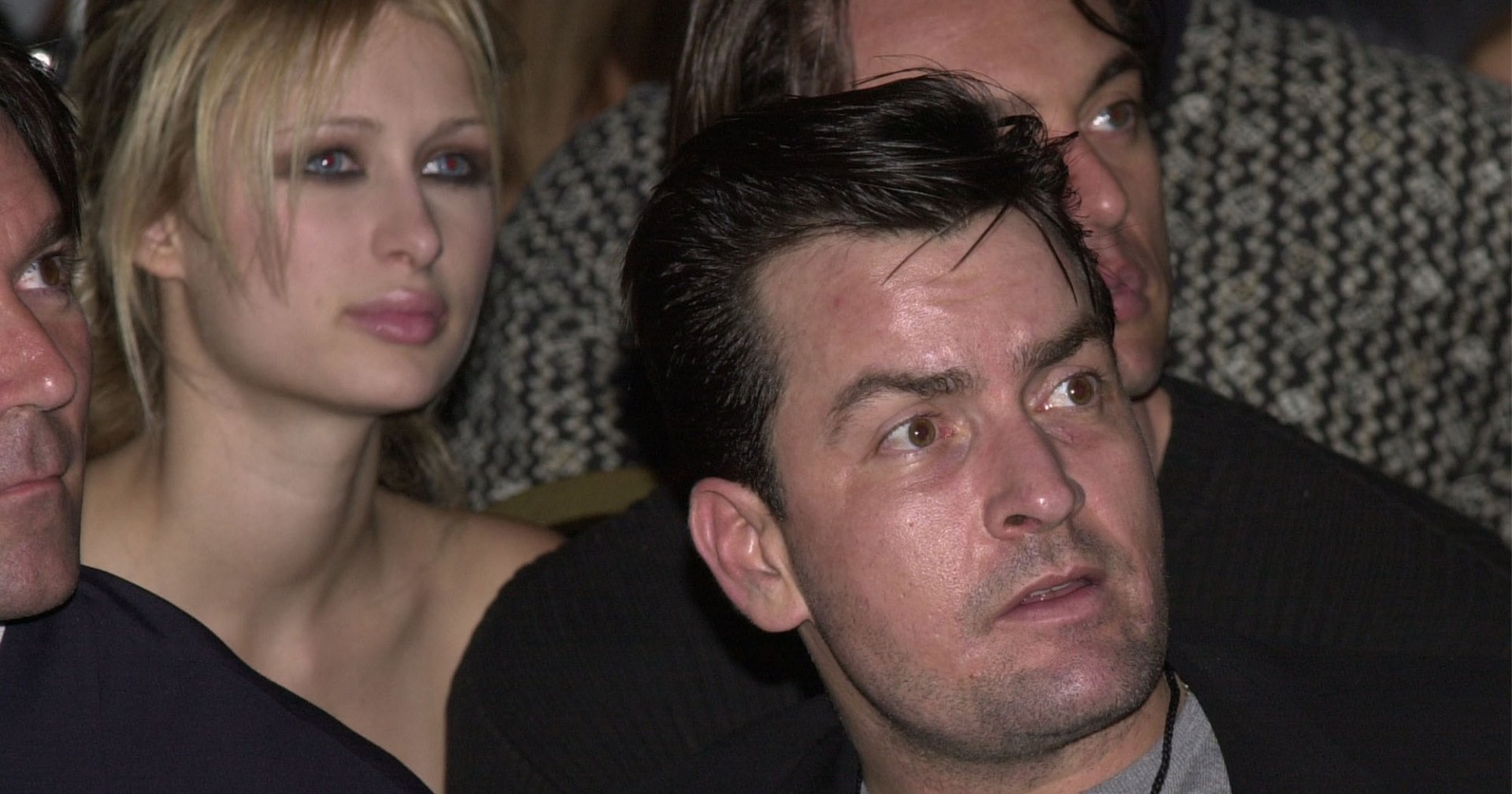 A dark-haired man in a blazer and grey turtleneck sits next to a blonde woman in a black dress at an indoor event.
