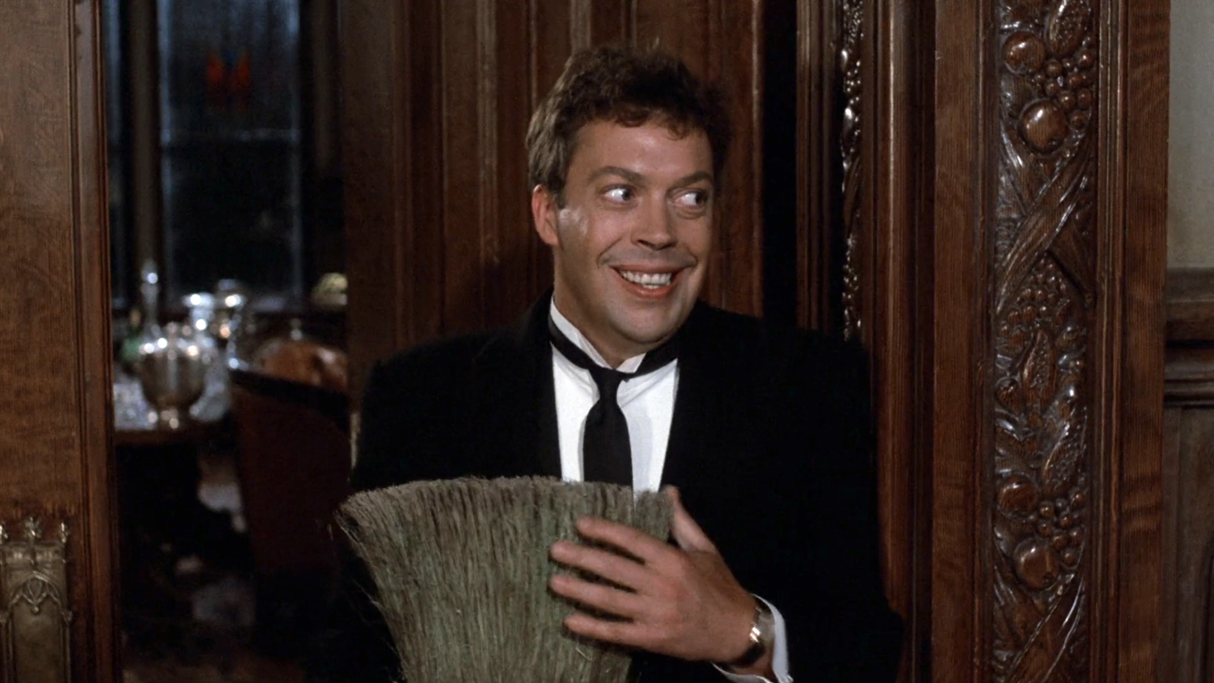 Man in black suit and tie holding a broom, smiling mischievously while glancing sideways through a carved wooden doorframe.