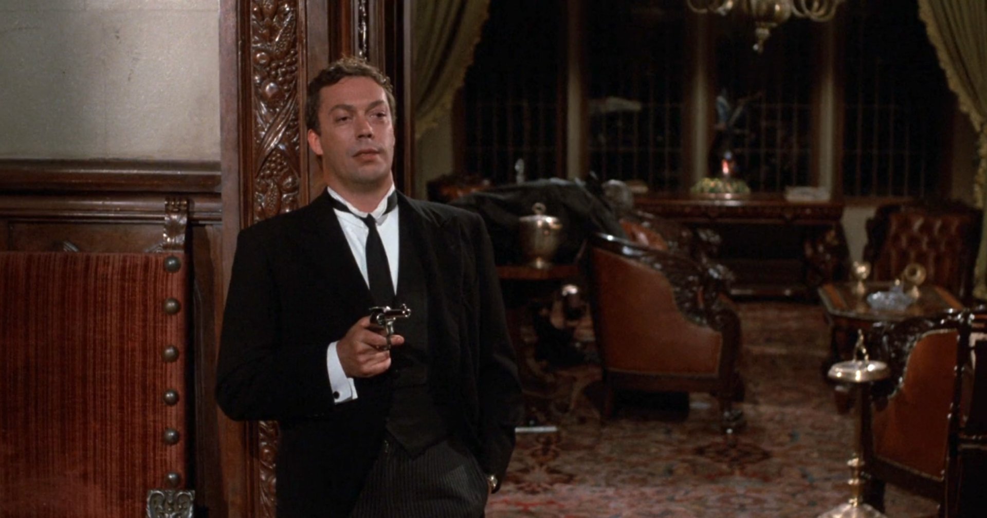 Man in black suit and tie holding a broom, smiling mischievously inside an ornate wood-paneled room, in a still from Clue (1985).
