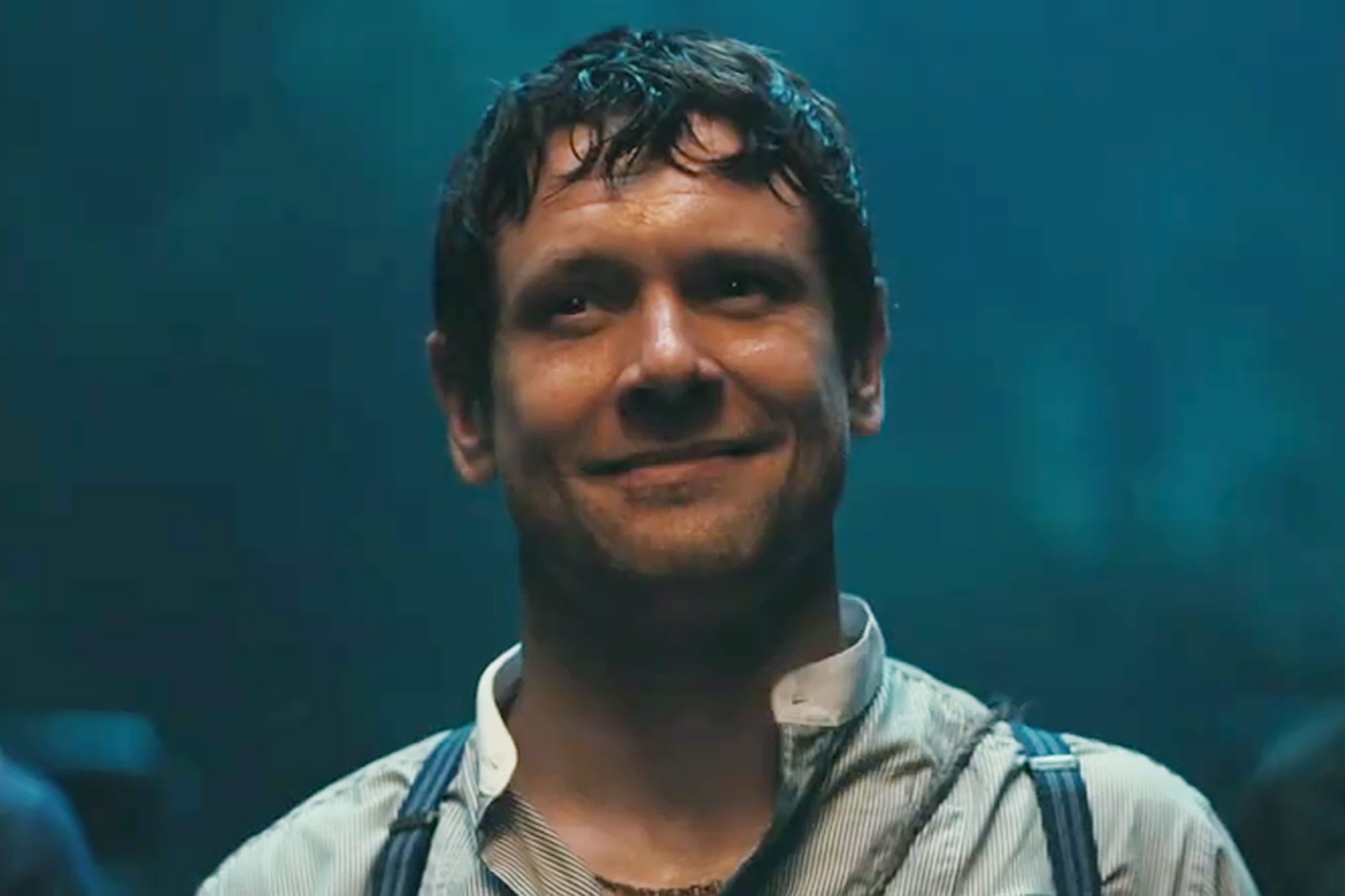 Man with dark curly hair smiling, wearing a striped shirt and suspenders in a blue-toned cinematic scene.