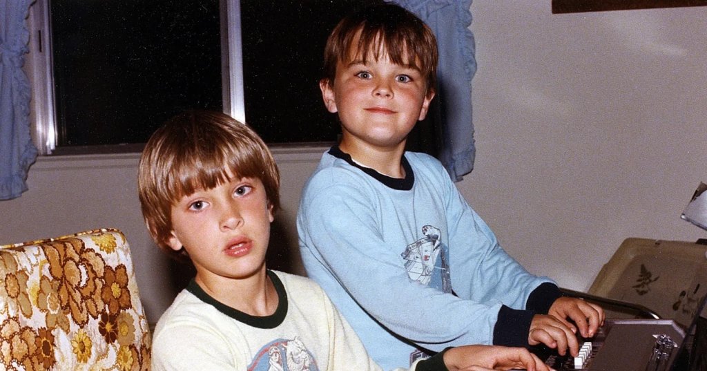 Two young boys in matching Star Wars pajamas sitting at a wooden keyboard organ in a home living room, vintage photo.