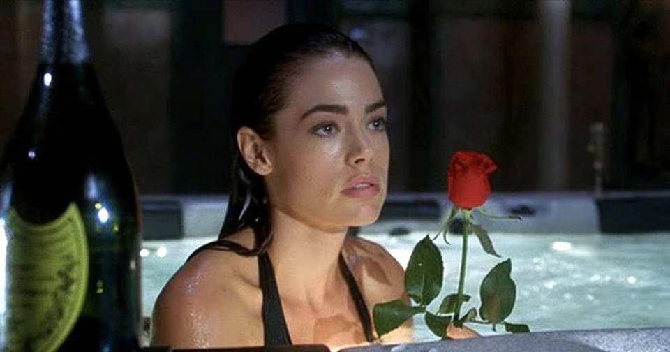 Woman in a black swimsuit sitting in a hot tub next to a champagne bottle and a red rose, looking off to the side.