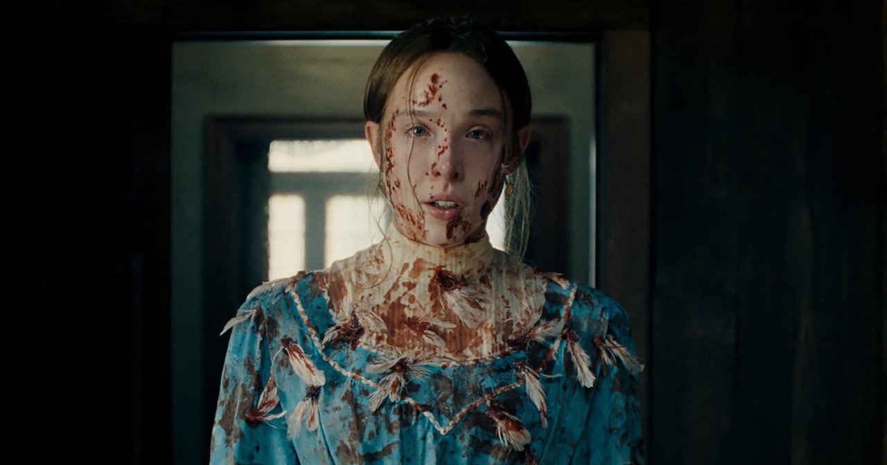 A young woman covered in blood stains on her face and blue Victorian dress stands in a dark doorway in a horror film still.