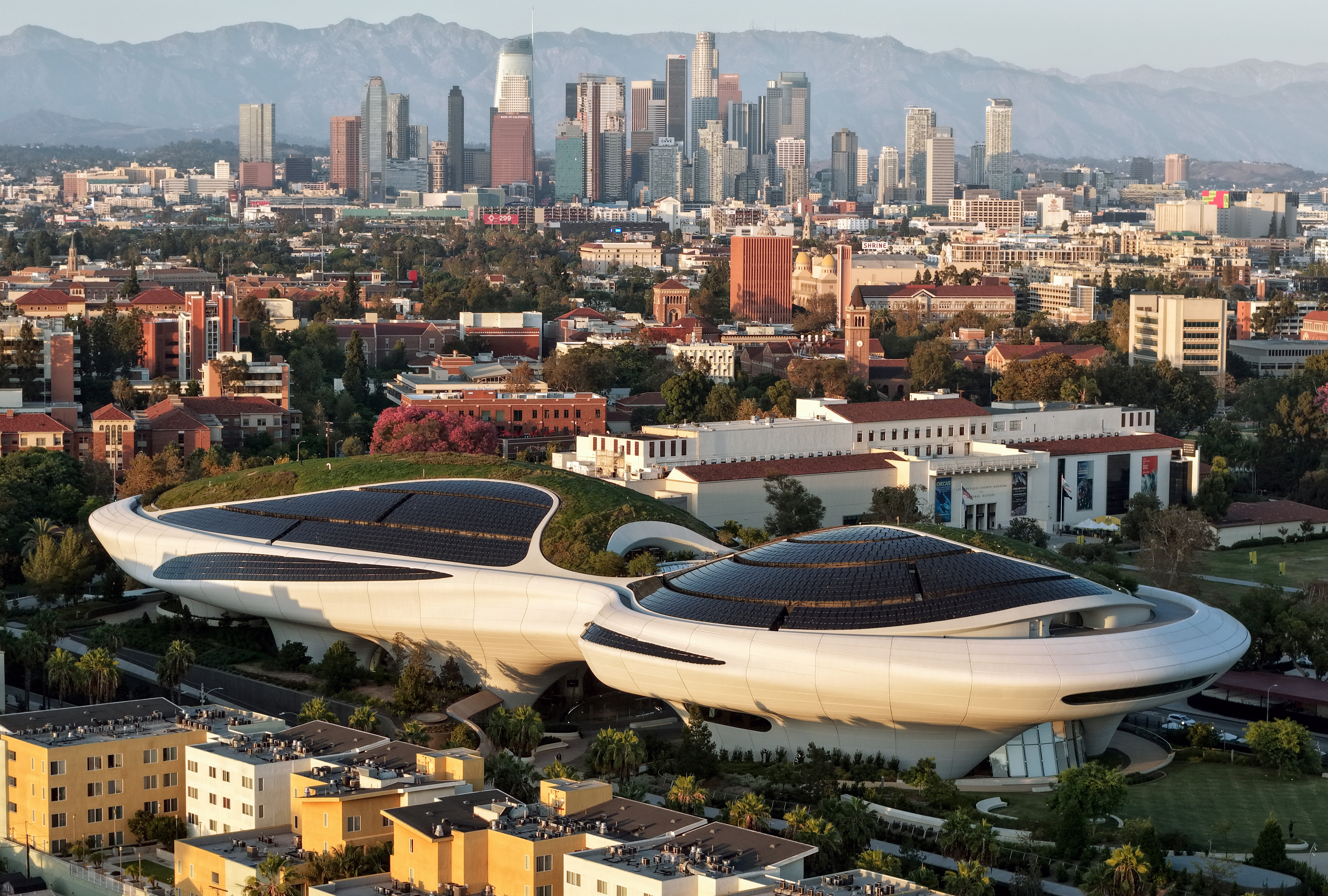 Aerial view of the futuristic, curved Lucas Museum of Narrative Art with solar panels on its roof, set against the Los Angeles skyline and mountains.