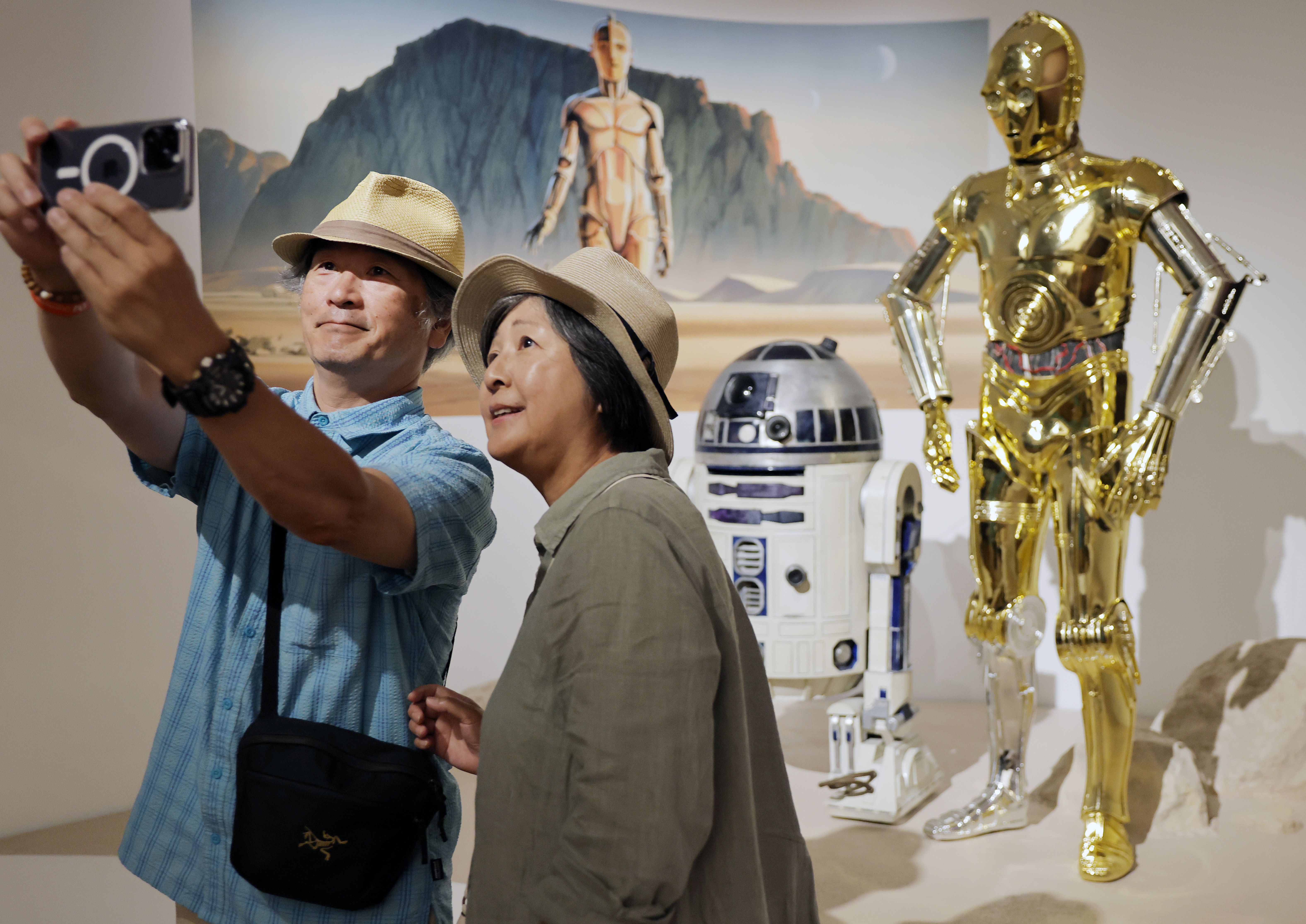 Two museum visitors in straw hats take a selfie in front of R2-D2 and C-3PO props inside a Star Wars exhibit.