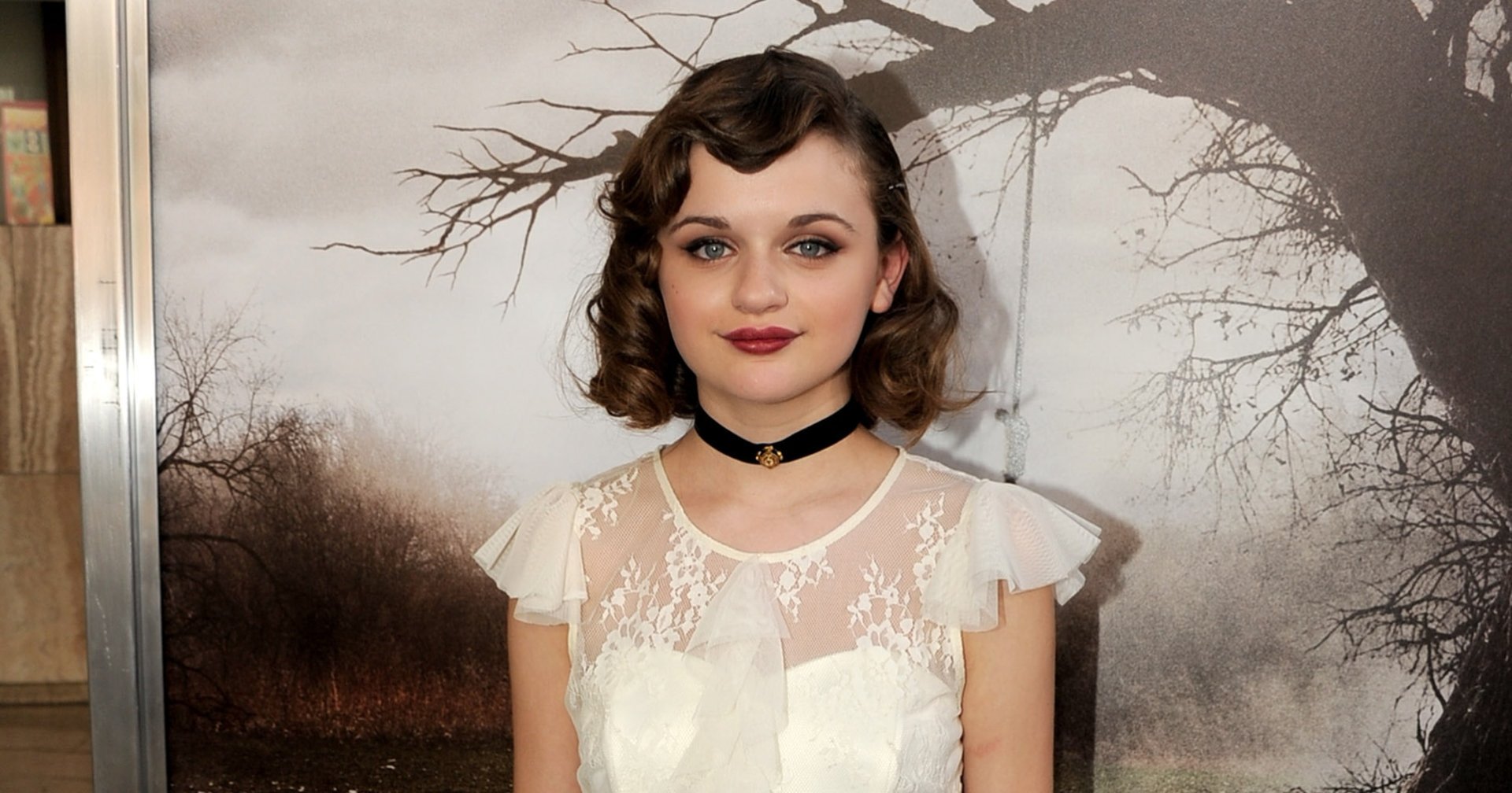 Joey King wearing a white tiered lace dress and black choker, posing in front of The Conjuring movie premiere backdrop at the Cinerama Dome, Los Angeles.