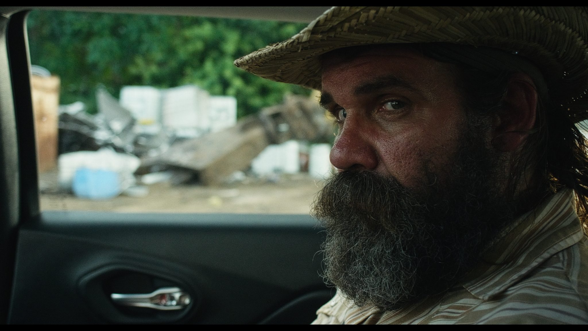 Bearded man in a straw hat and striped shirt sitting in a car, looking to the side, with a cluttered junkyard visible through the rear window.
