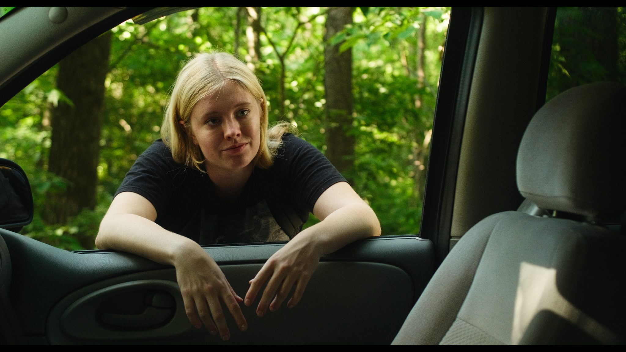 Young blonde woman in a black t-shirt leaning arms-first through an open car window, green wooded background behind her.