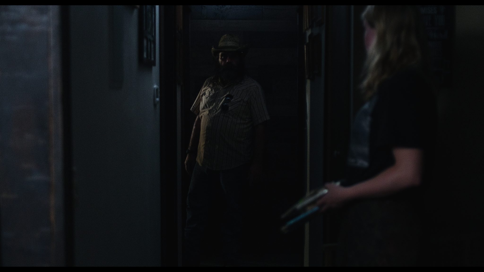 Dimly lit corridor scene with a man in a straw hat standing in a shadowy doorway and a woman in the foreground holding an object.