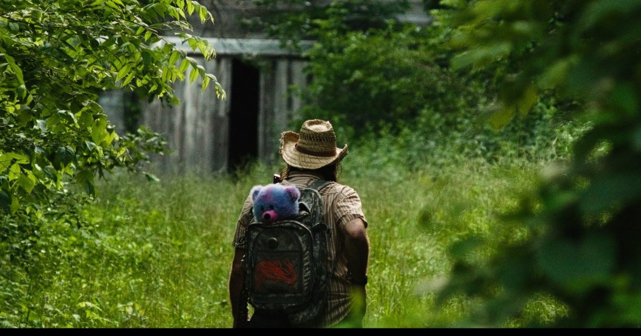 Man in straw hat with backpack carrying a stuffed animal walks through tall grass toward a dilapidated wooden barn surrounded by lush greenery.