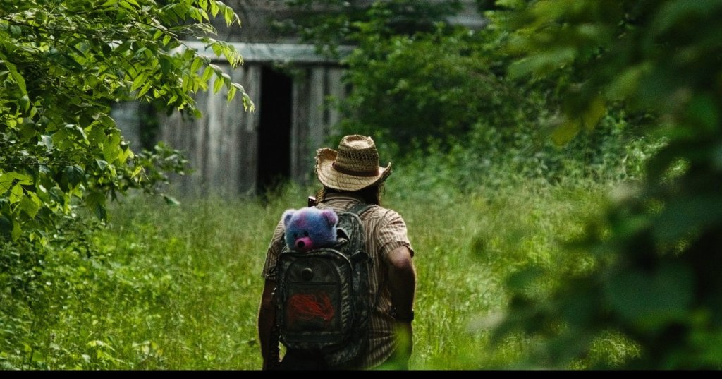 Man in straw hat with backpack carrying a stuffed animal walks through tall grass toward a dilapidated wooden barn surrounded by lush greenery.