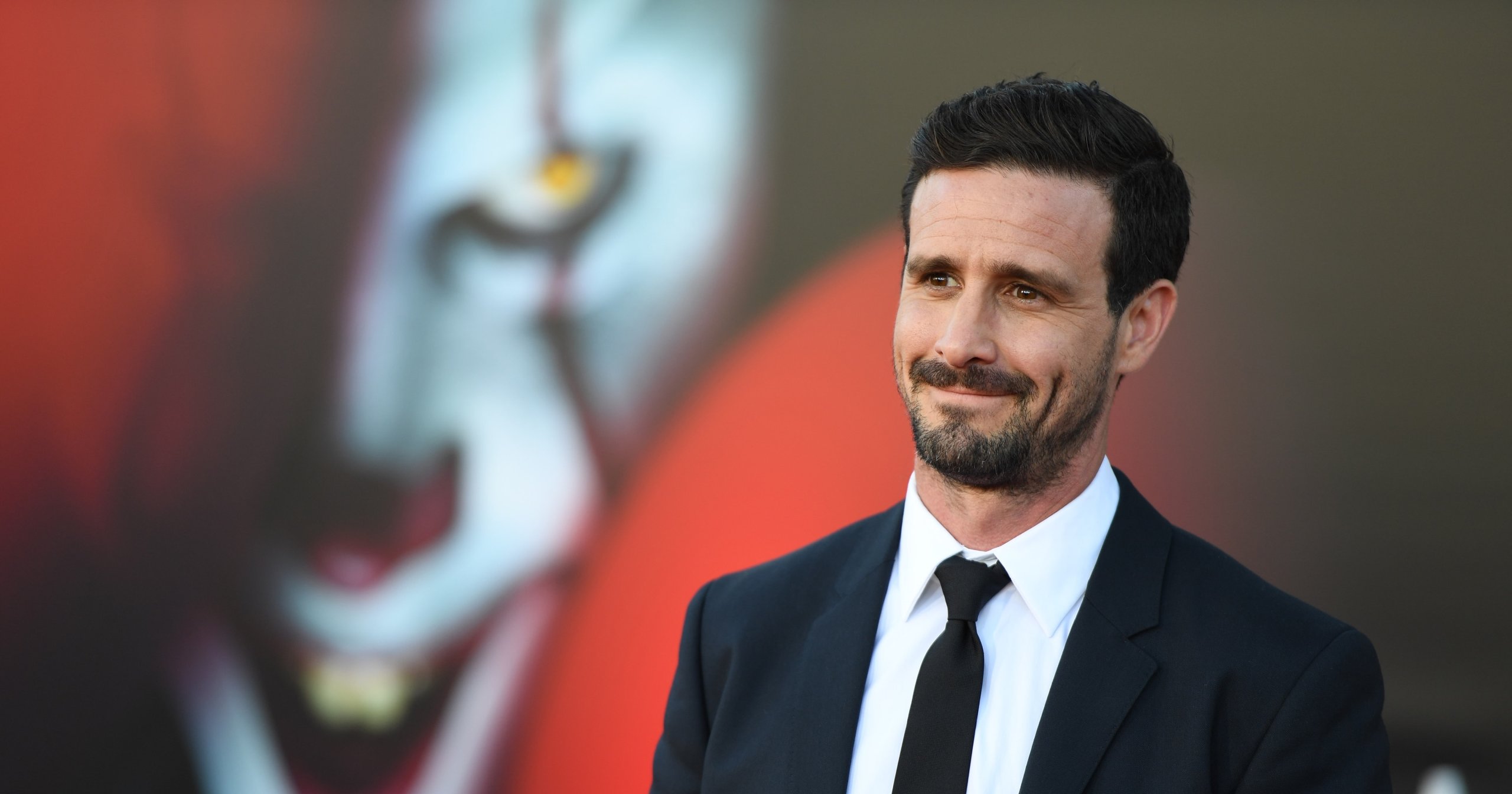 Man in black suit and tie smiling at a premiere, with a blurred clown image in the background.