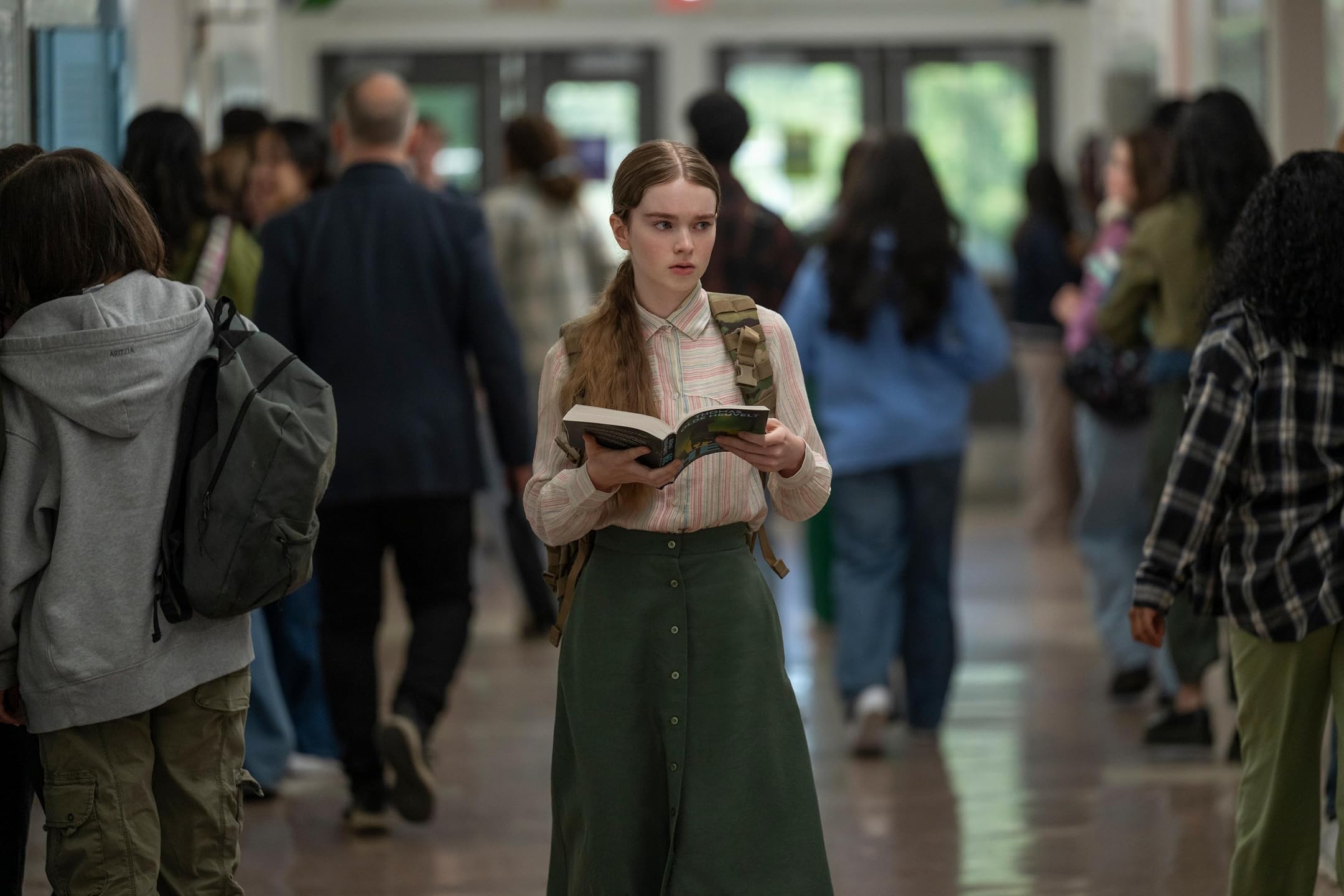 A teenage girl with long braided hair in a striped blouse and green maxi skirt holds an open book while walking through a busy school hallway.