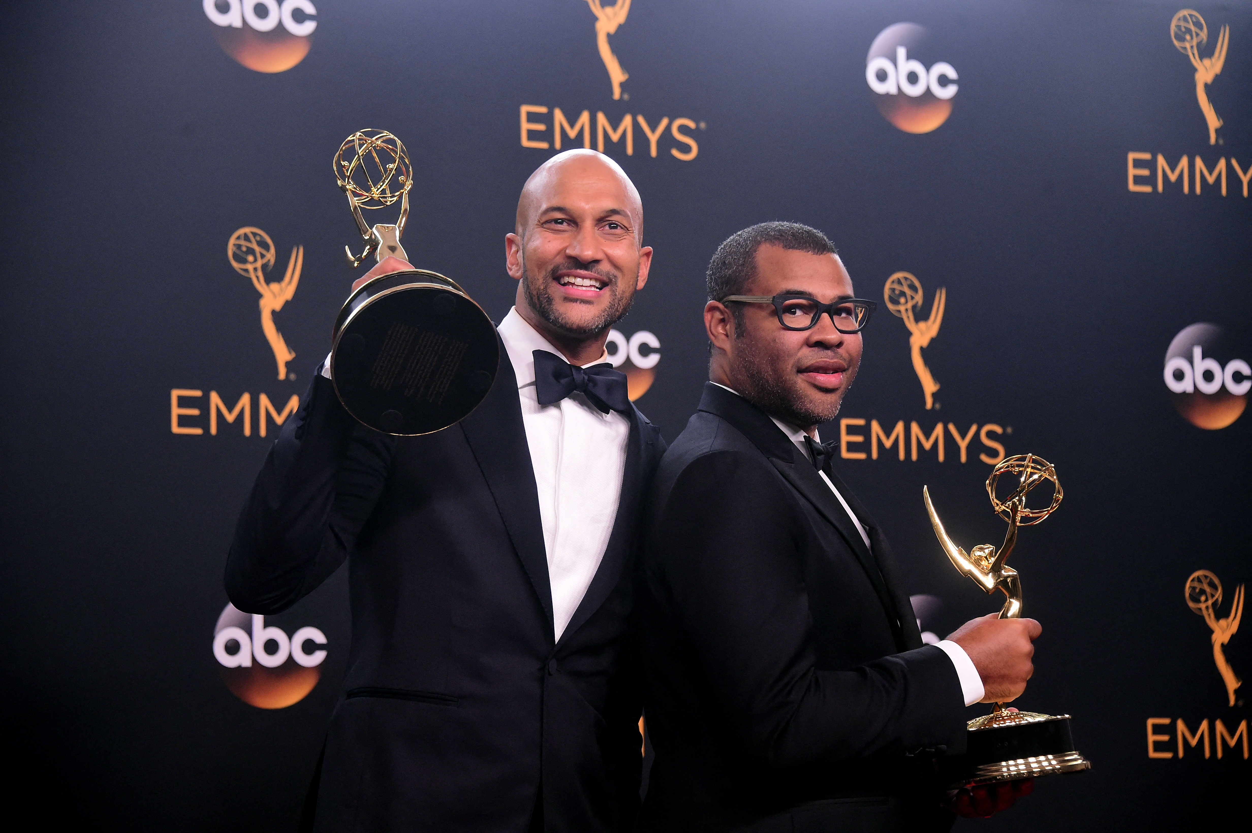Two men in tuxedos holding Emmy Award statuettes and posing back-to-back in front of an Emmys step-and-repeat backdrop.