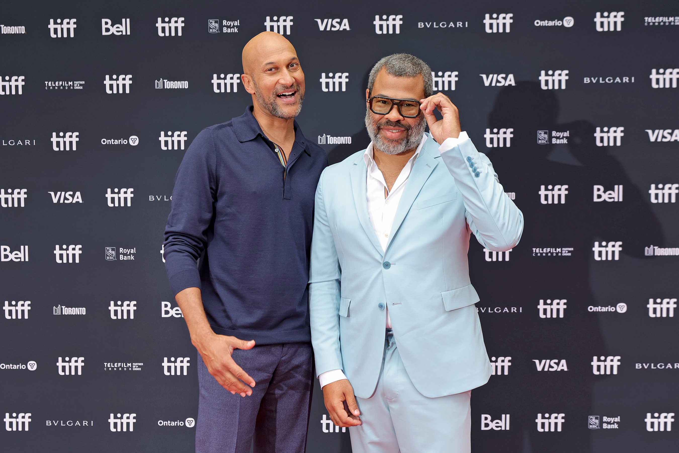 Two smiling men standing in front of a TIFF step-and-repeat backdrop, one in a navy polo shirt and one in a light blue suit adjusting his dark-framed glasses.