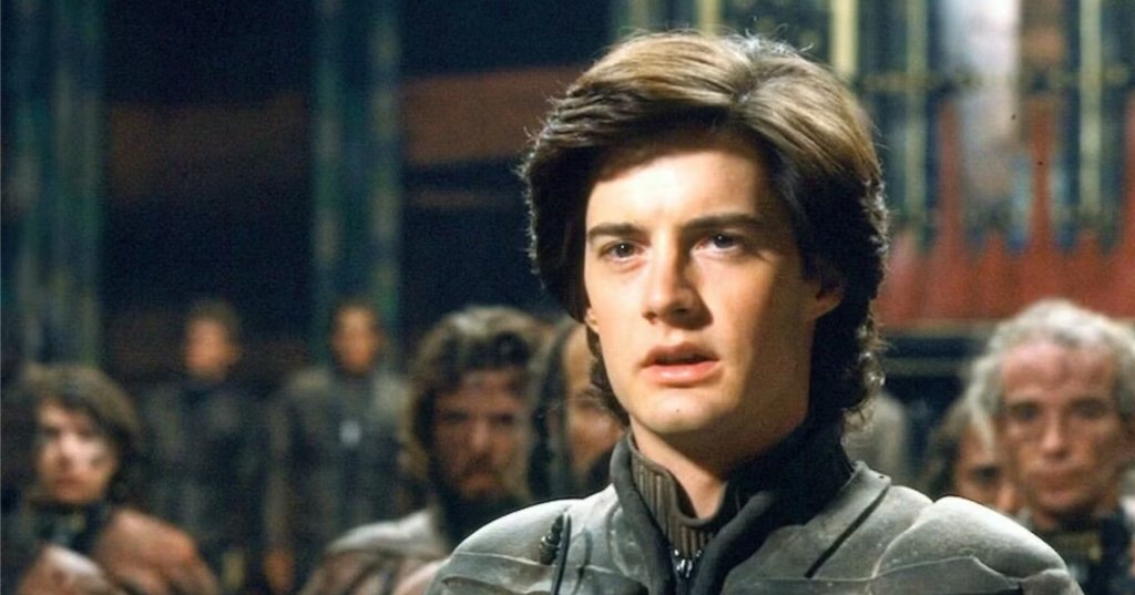 Young man in gray futuristic armor looking up with an intense expression, crowd of people blurred in the background, film still from Dune (1984).