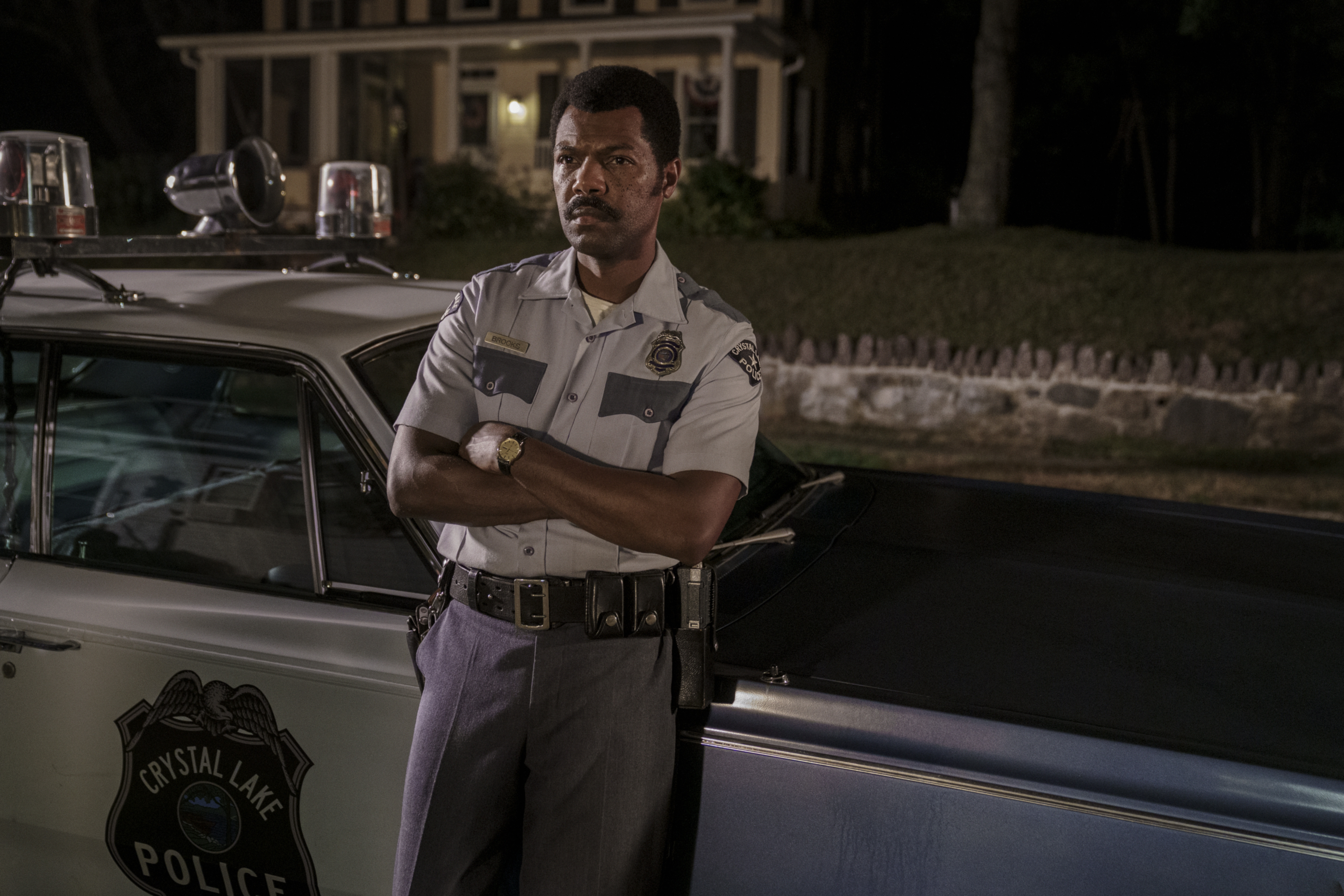 A man in a Crystal Lake Police uniform stands with arms crossed beside a vintage police car at night.