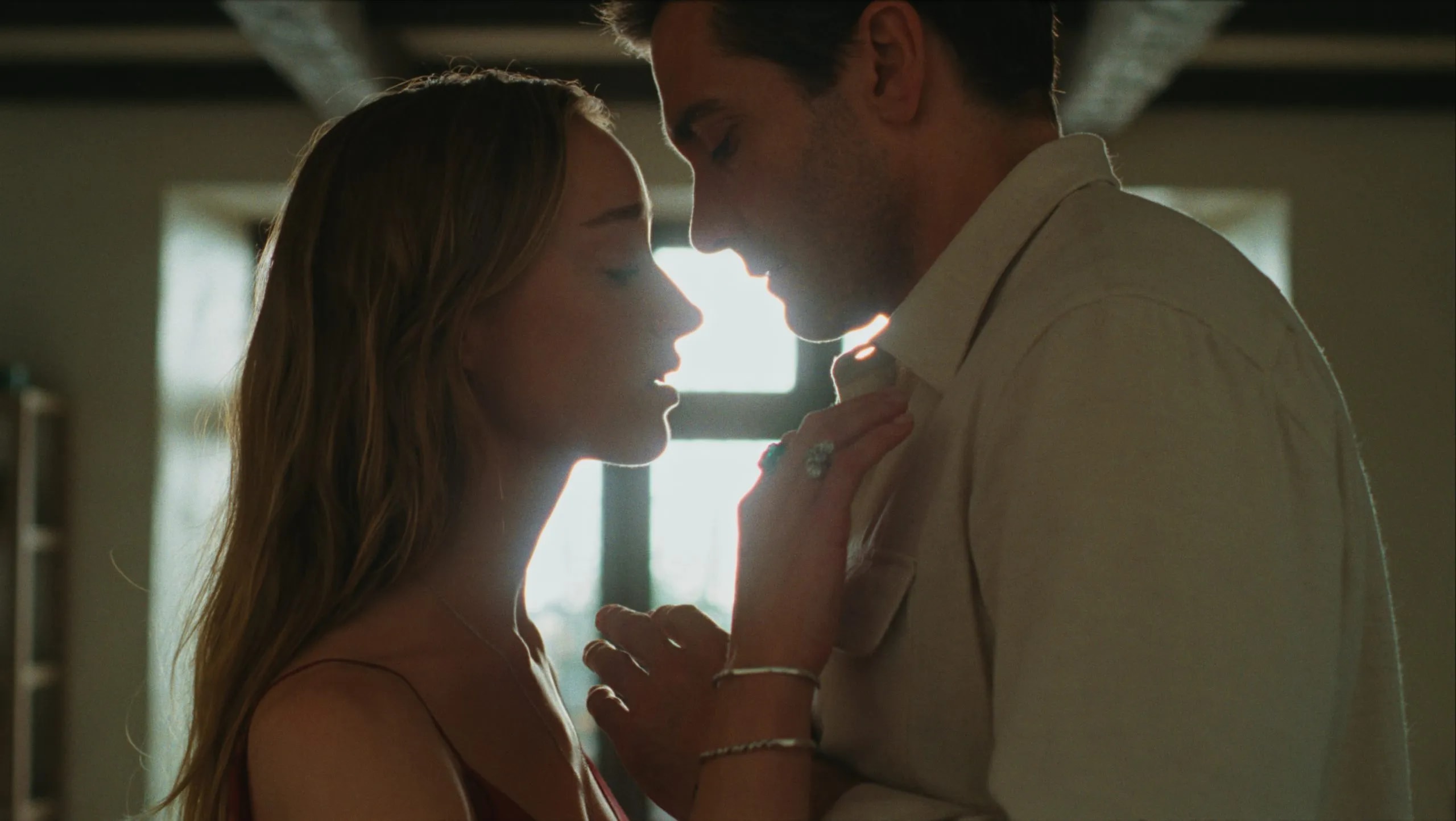 A man and woman lean their faces close together in a dimly lit room, her hands resting on his chest, backlit by bright window light.