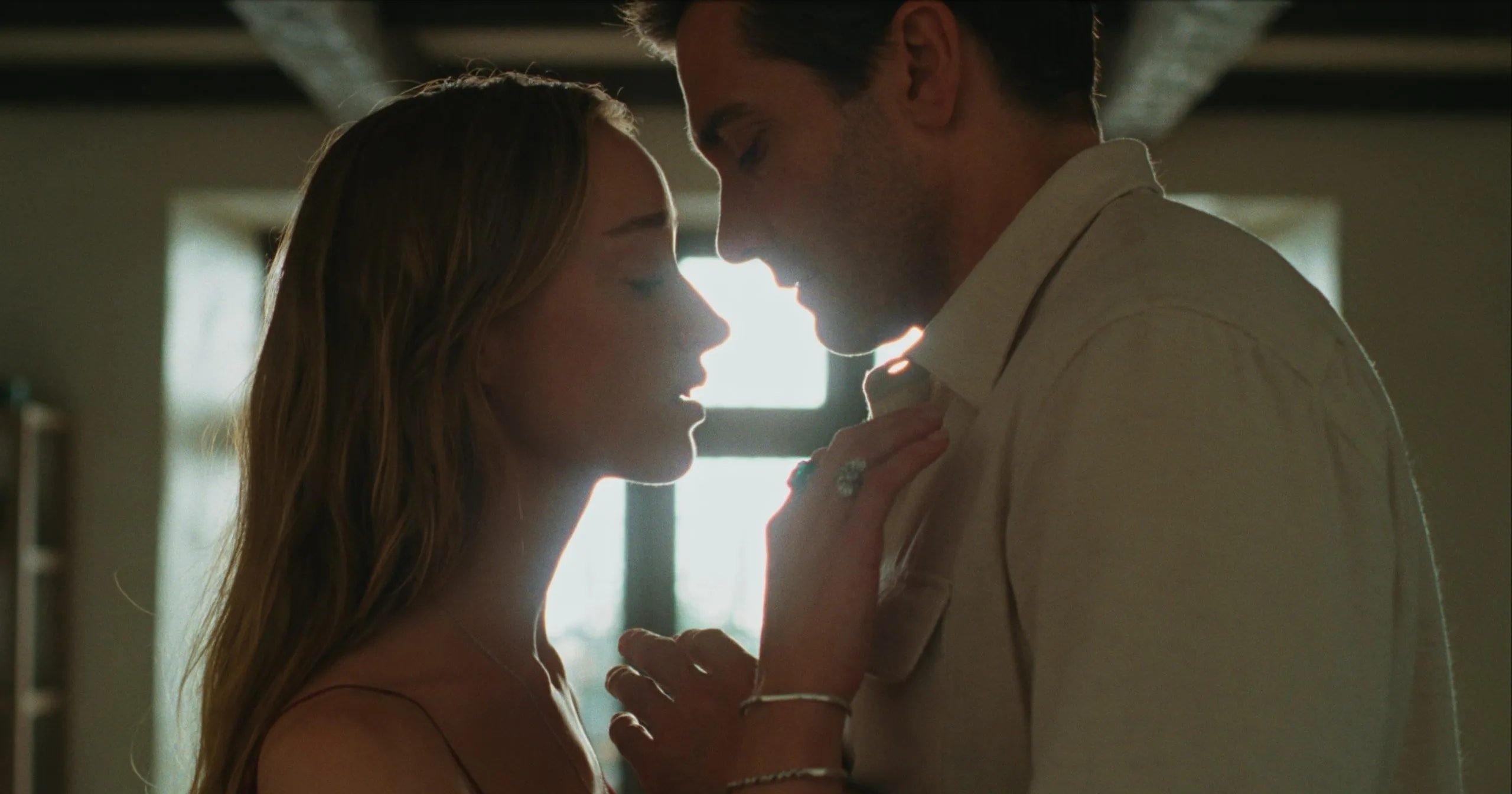 A woman in a red dress and a man in a white shirt lean in close, faces nearly touching, bathed in soft backlight indoors.