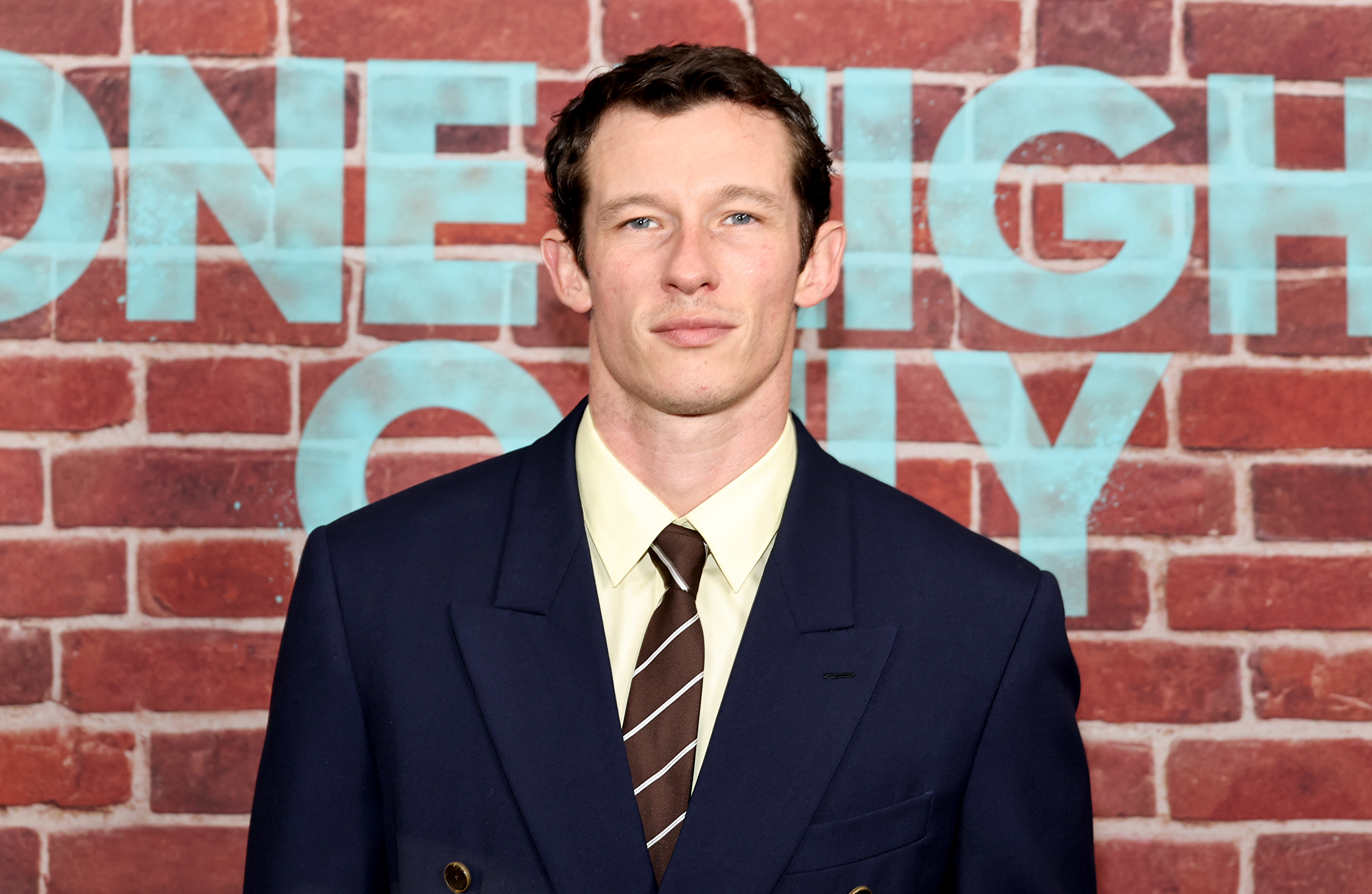 Callum Turner in a navy double-breasted suit and striped tie stands in front of a brick wall with "One Night Only" text at SVA Theater, NYC.