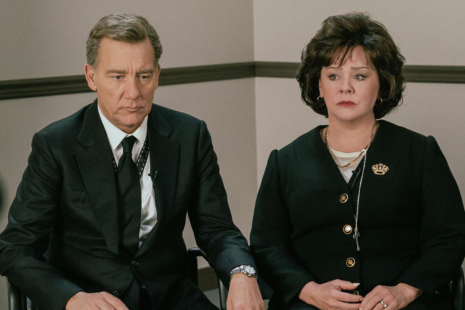 A man in a dark suit and black tie sits beside a woman in a black blazer with a gold brooch, both looking tense and somber.