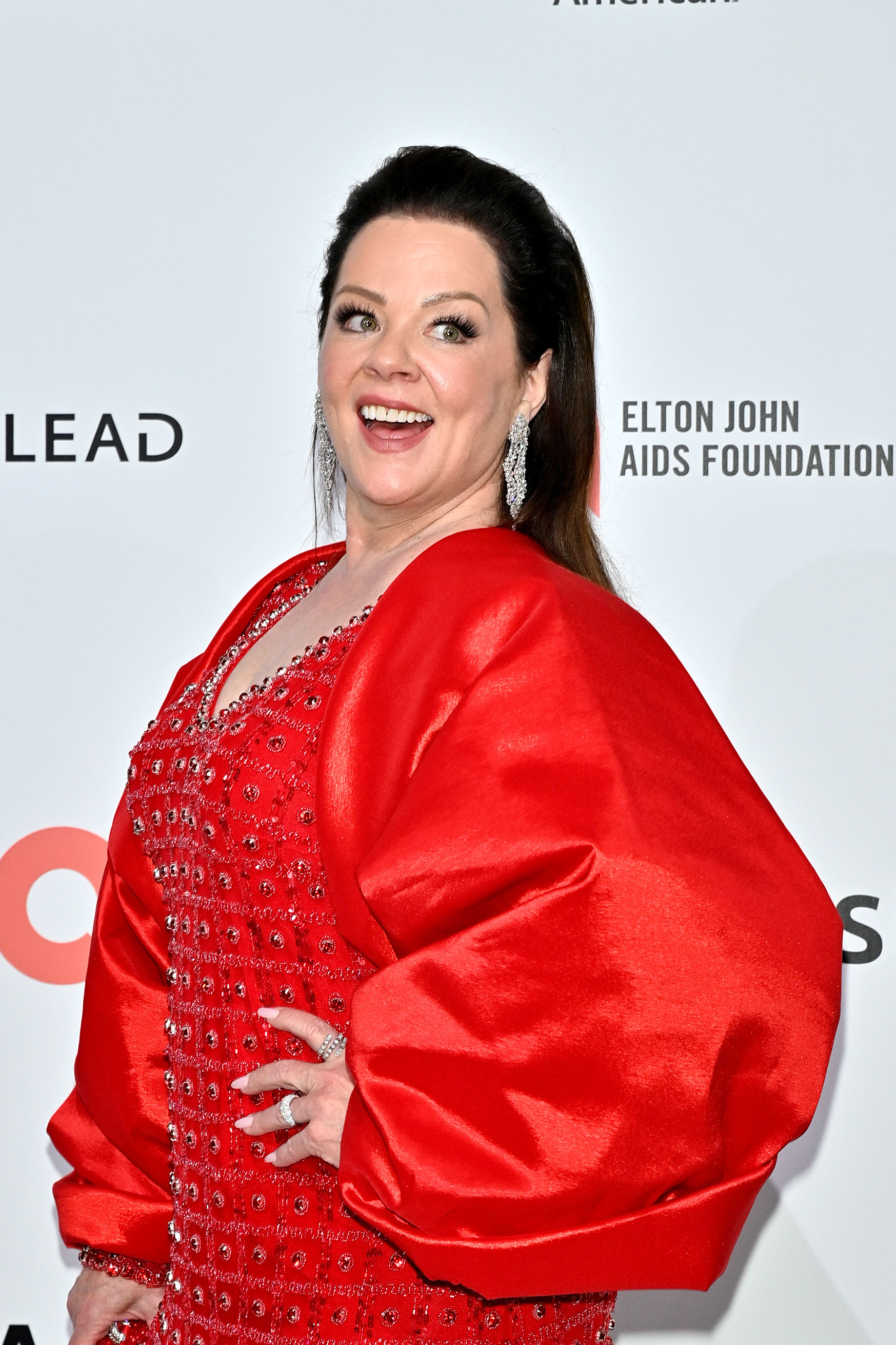 Melissa McCarthy in a red embellished gown with dramatic satin sleeves smiles at the Elton John AIDS Foundation Oscar viewing party step-and-repeat.