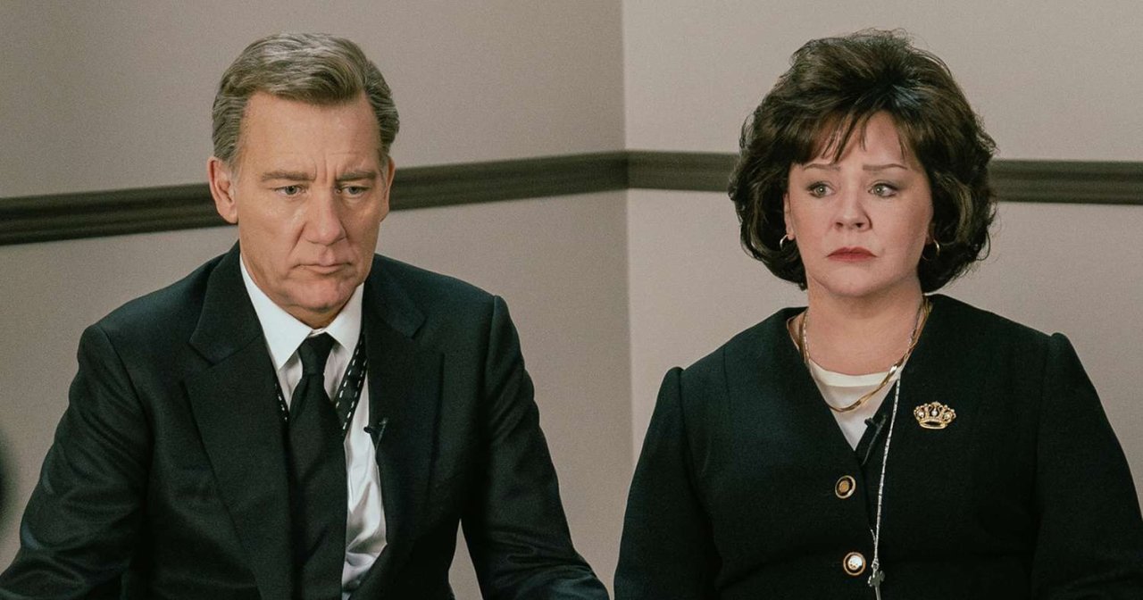 A man in a black suit and tie and a woman in a dark blazer with a gold brooch sit together looking somber in a TV studio scene.