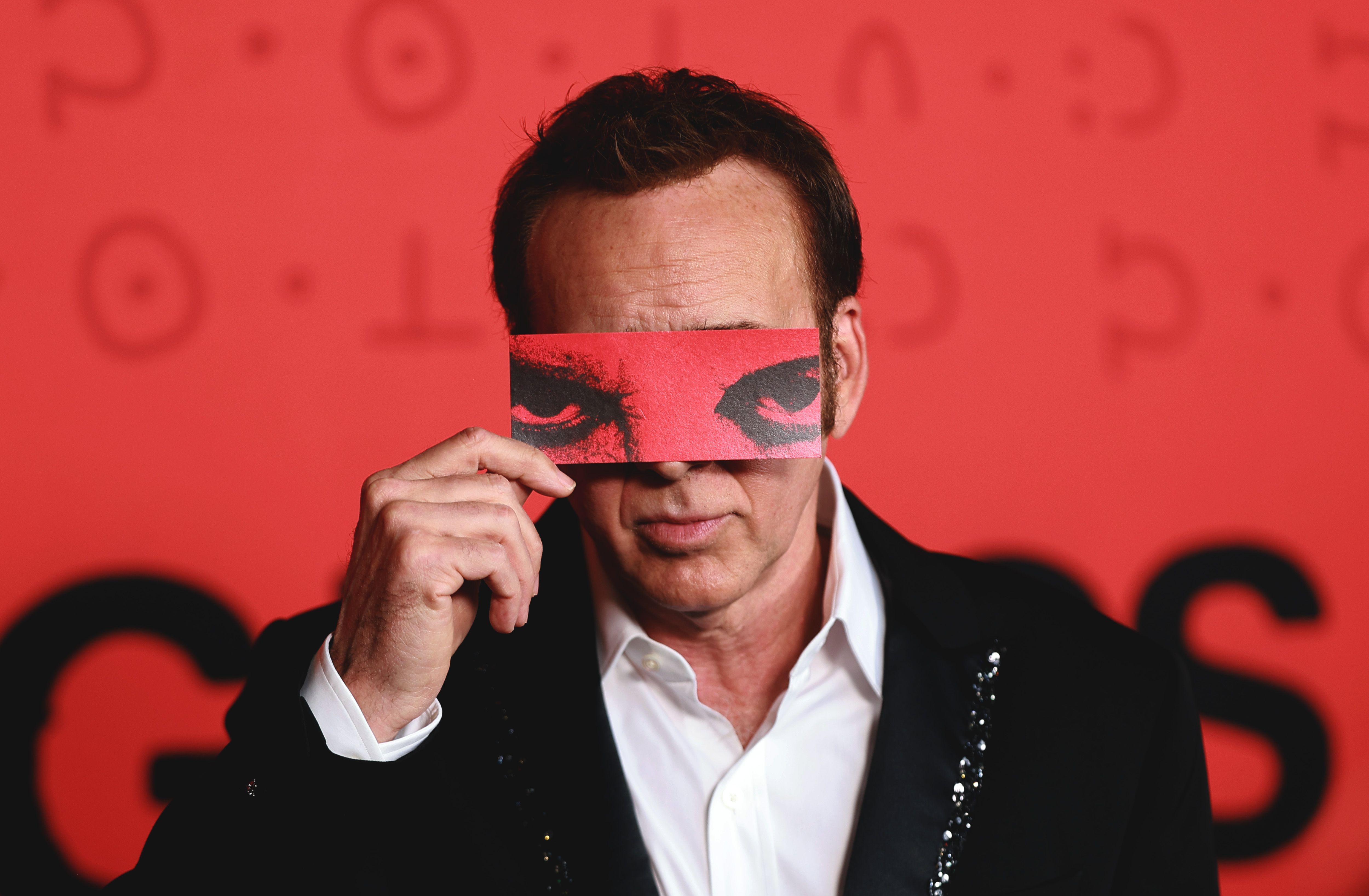 Man in black sequined blazer holds a red card printed with menacing eyes over his face in front of a red backdrop.