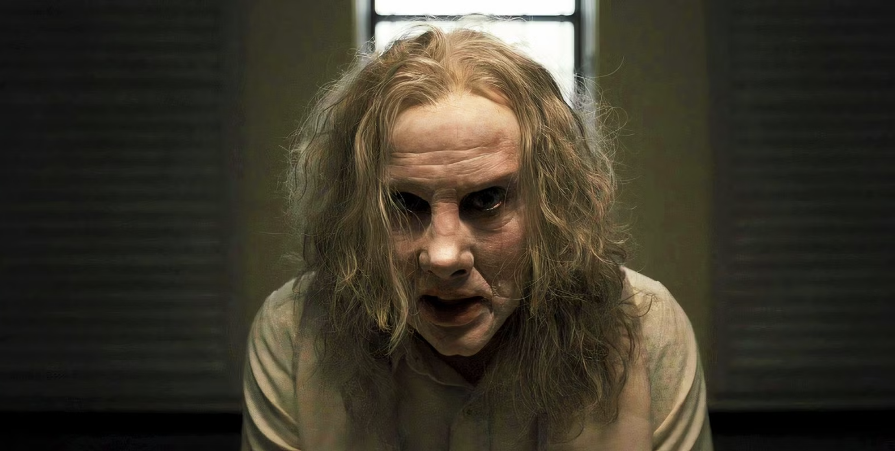 A gaunt, pale figure with long disheveled hair and dark sunken eyes leans toward the camera in a dimly lit room.