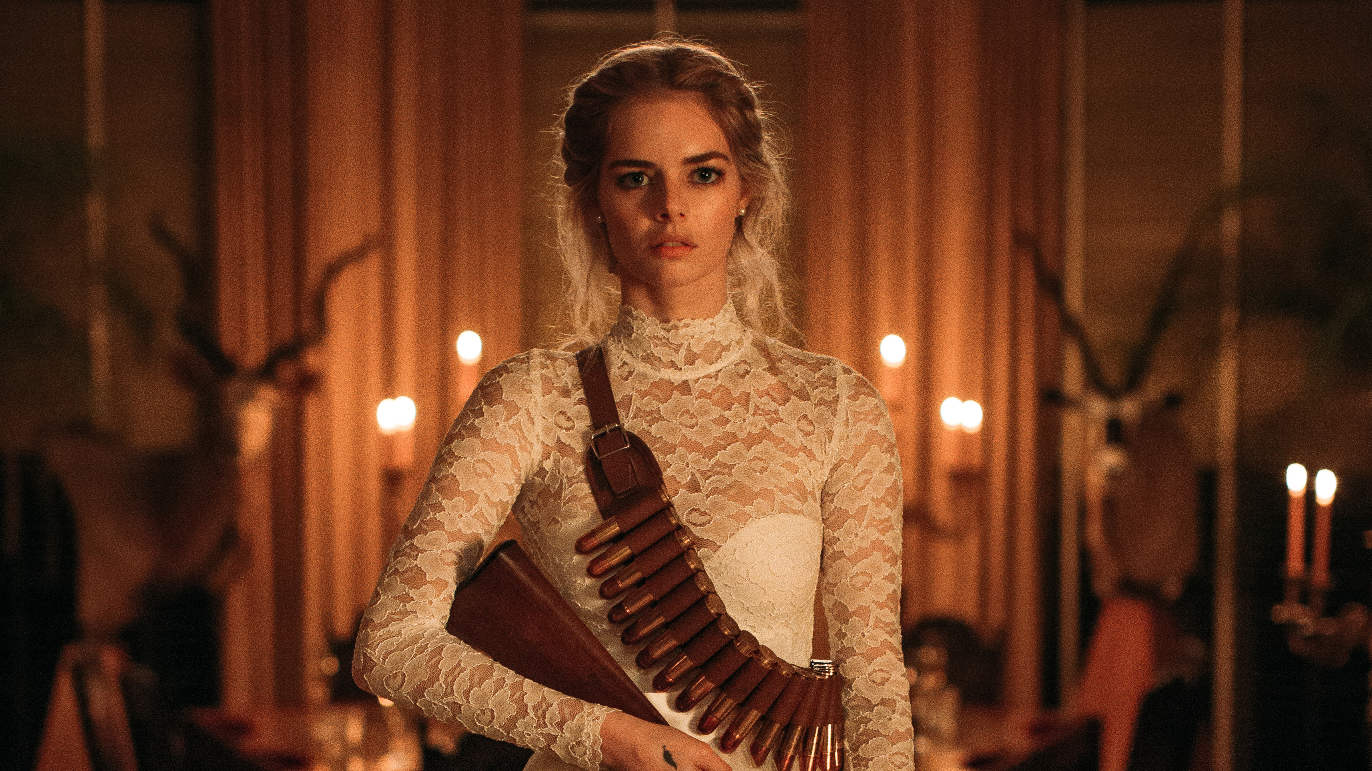 A blonde woman in a white lace dress wearing a leather bullet bandolier stands in a dimly candlelit room with animal mounts on the walls.
