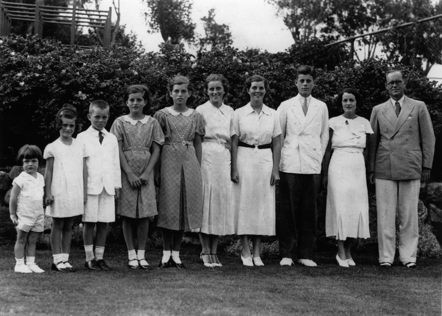 Black-and-white outdoor photo of the Kennedy family standing in a row on a lawn in front of hedges, including parents and eight children, 1937.