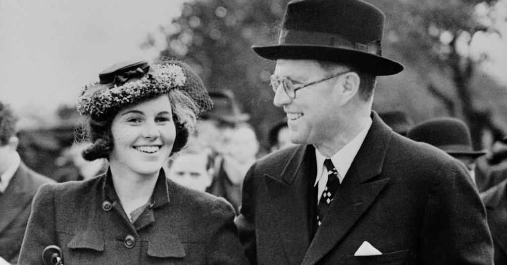 Black-and-white photo of a young smiling woman in a coat and floral hat walking arm-in-arm with an older man in a suit and fedora, 1938.