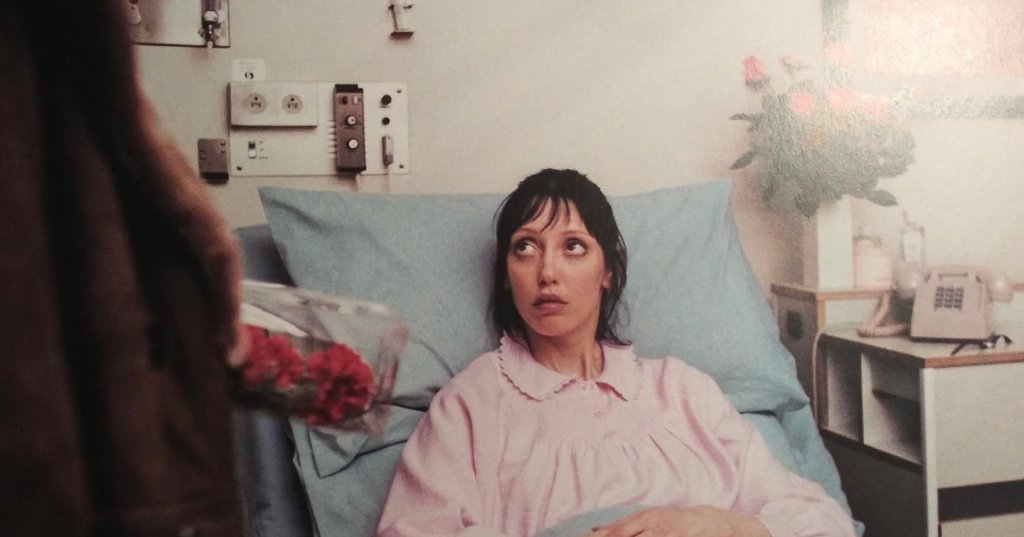 Woman in pink hospital gown lying in a hospital bed looking up nervously while someone offers her red flowers, from The Shining (1980).