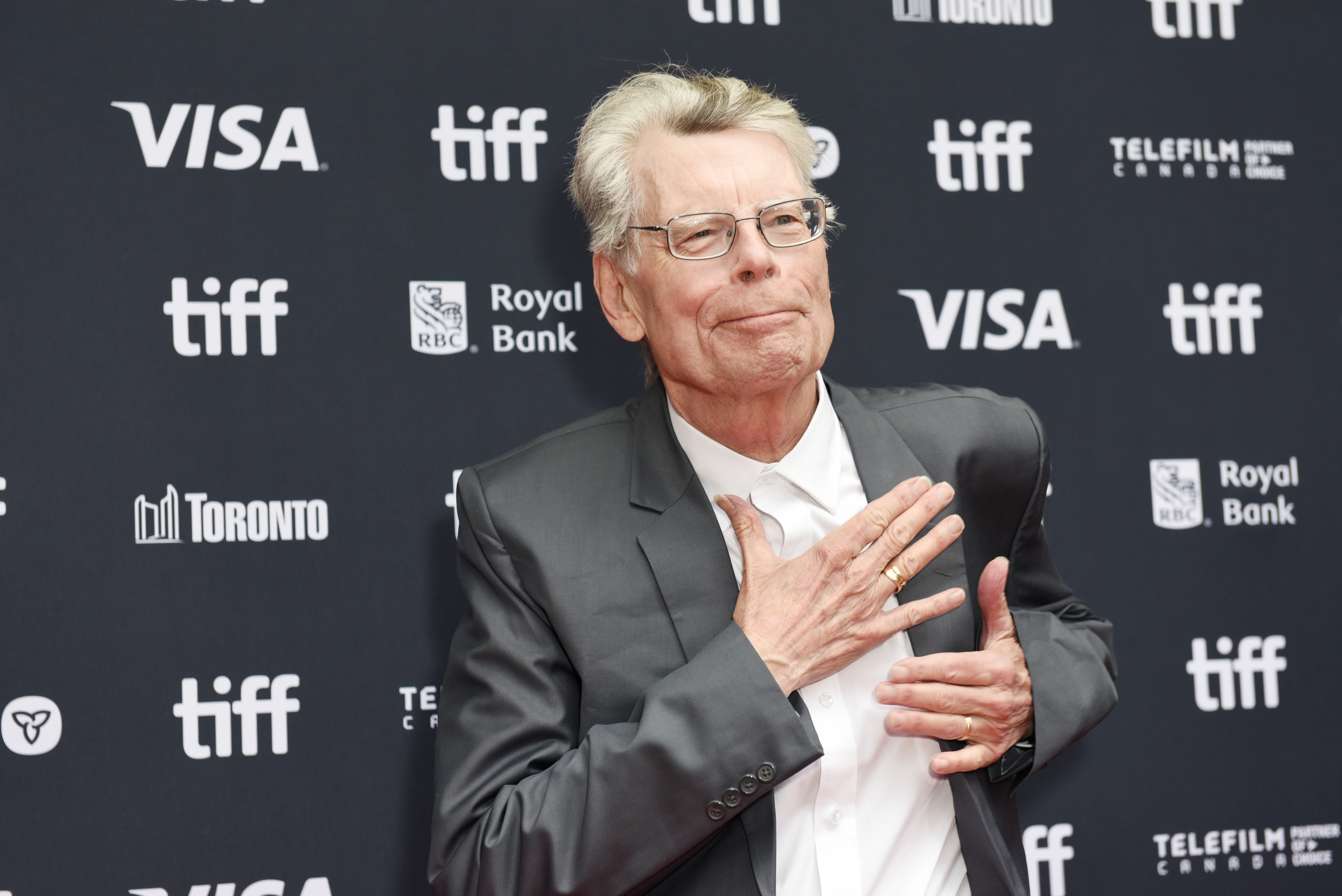Stephen King in a dark suit gesturing at his chest on the TIFF red carpet, with Visa, Royal Bank, and Toronto sponsor logos behind him.
