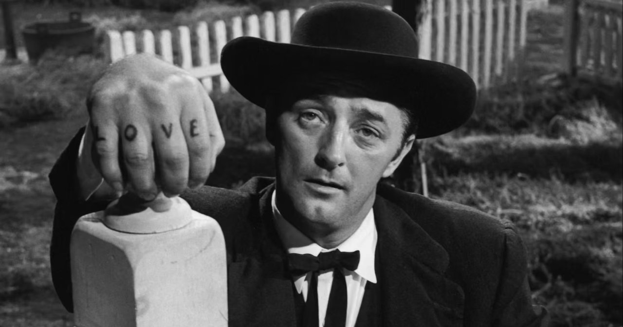 Black-and-white film still of a man in a wide-brimmed hat resting a fist tattooed with "LOVE" on a gravestone, staring intensely.