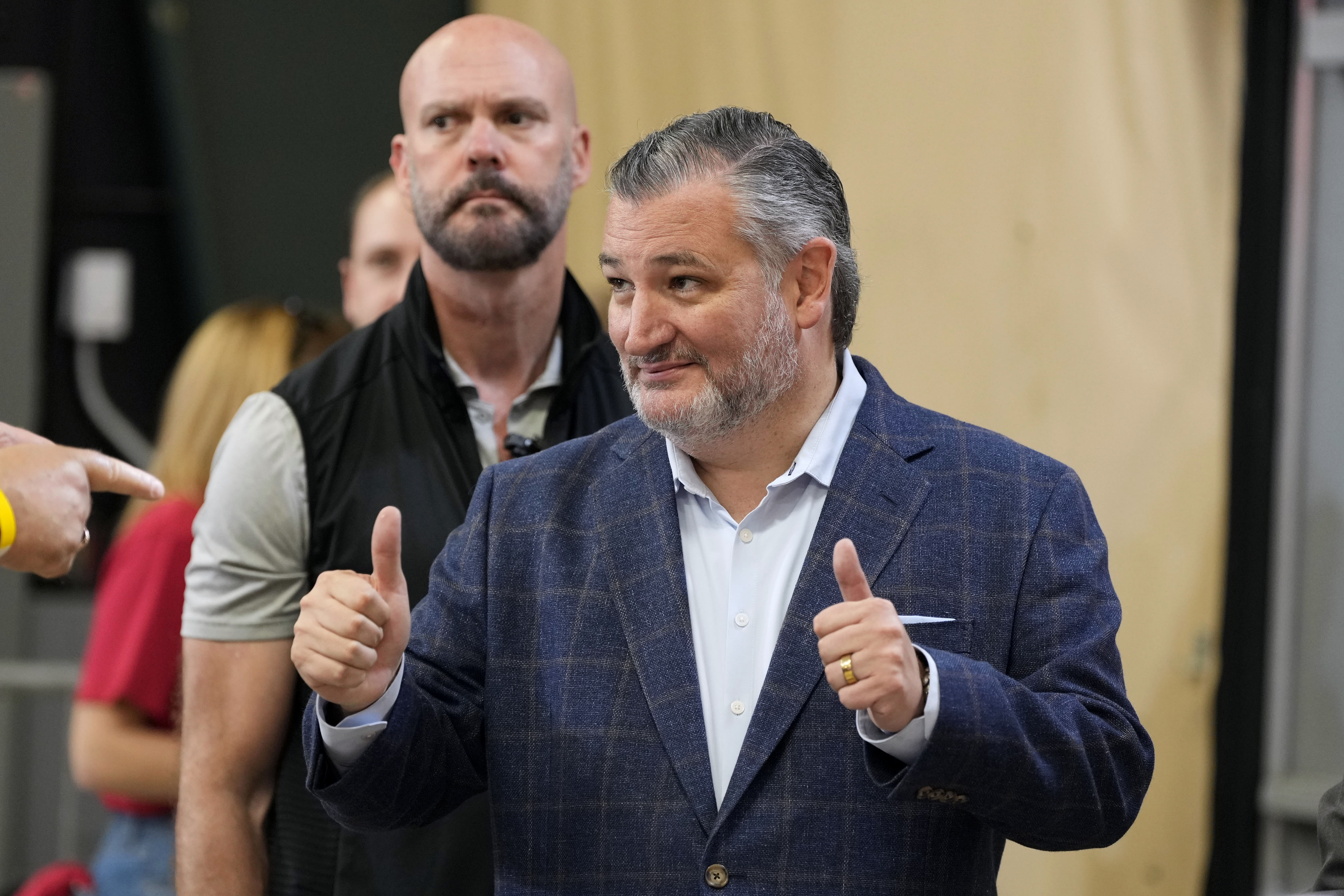 Sen. Ted Cruz giving two thumbs up and smiling at an indoor campaign fundraiser event, with a bald man in a black vest standing behind him.