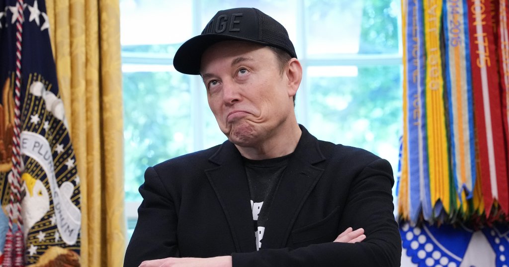 Elon Musk stands arms crossed in the Oval Office, wearing a black cap and blazer, with the presidential seal visible to his left.