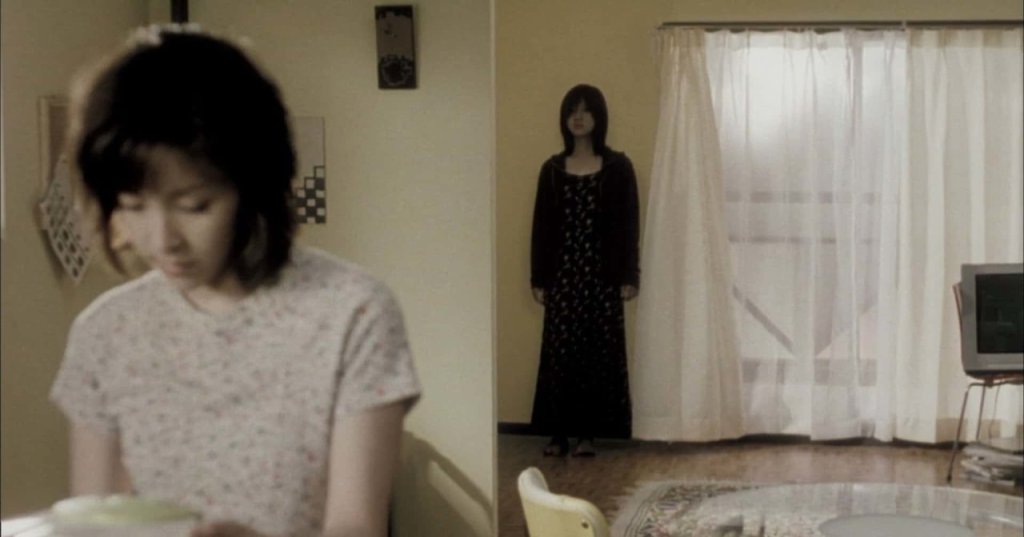 Split-focus shot: a woman in a floral shirt in the foreground and a dark-dressed figure standing still by sheer curtains in the background of a dim apartment.