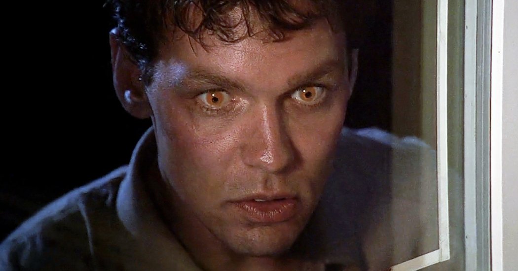 A man with glowing amber eyes and dark curly hair peers menacingly through a window frame against a dark background.