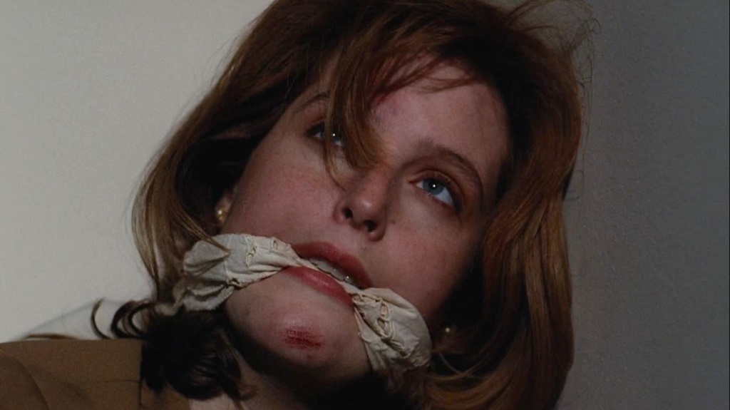A red-haired woman with blue eyes is bound and gagged with cloth around her mouth, looking upward with a distressed expression.