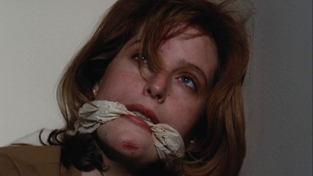 A red-haired woman with blue eyes is bound and gagged with cloth around her mouth, looking upward with a distressed expression.