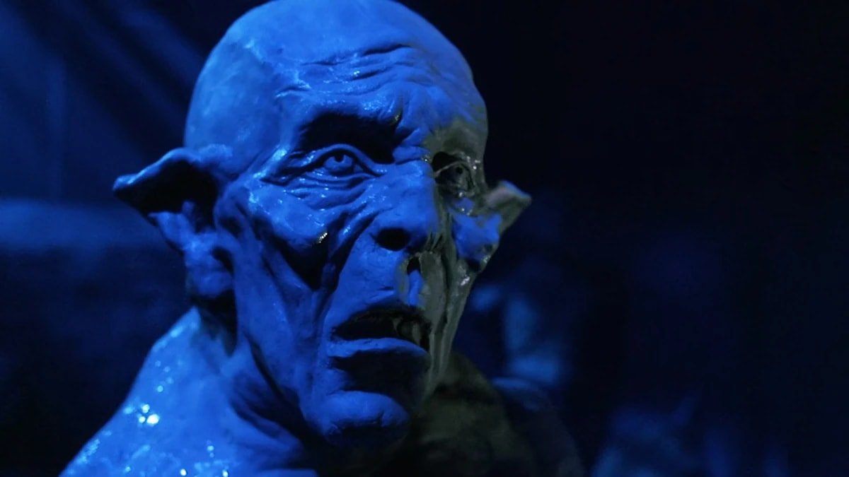 Close-up of a grotesque monster with wrinkled, decaying skin and pointed ears, lit by a harsh blue light against a dark background.