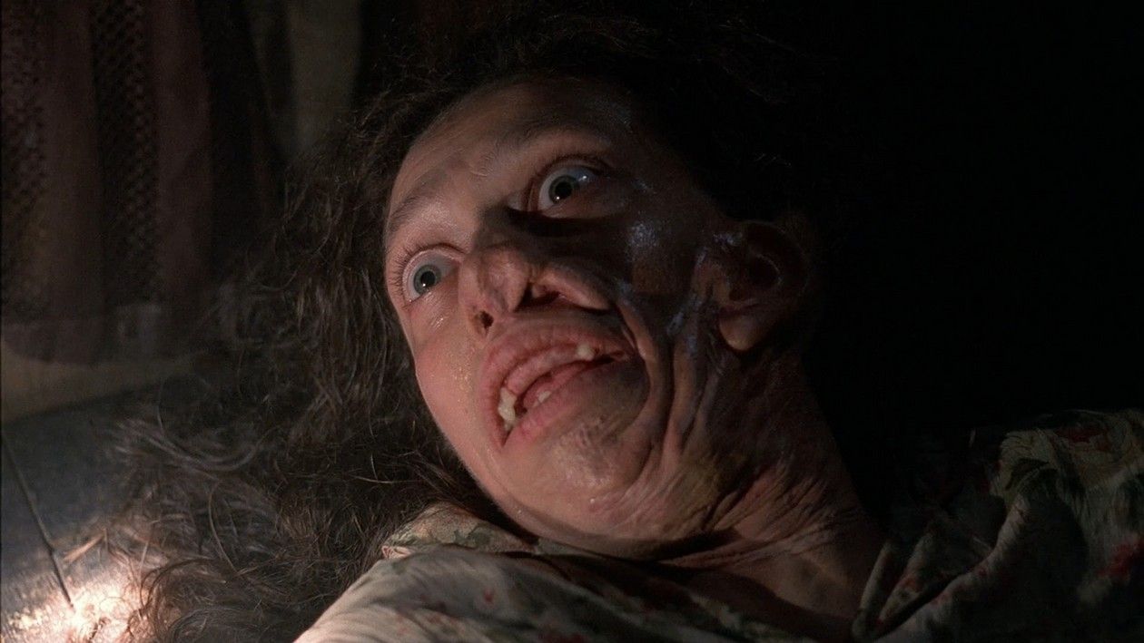 Close-up of a woman with heavily distorted, disfigured facial prosthetics lying down in a dark room, mouth agape.