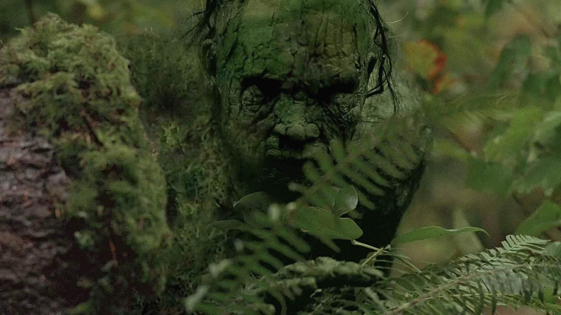 A green, heavily textured bark-like humanoid creature face partially hidden among lush ferns and forest foliage.