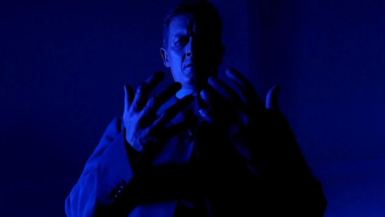 A man in a dark coat lit entirely in blue raises both hands with fingers spread wide, standing against a deep blue background.