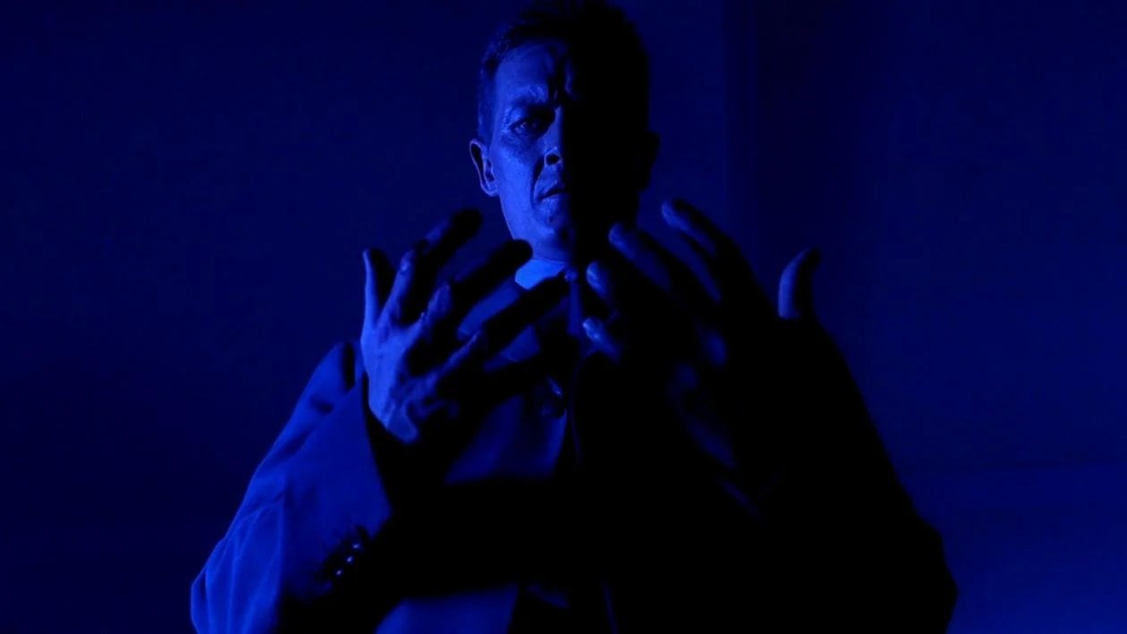 A man in a dark coat lit entirely in blue raises both hands with fingers spread wide, standing against a deep blue background.