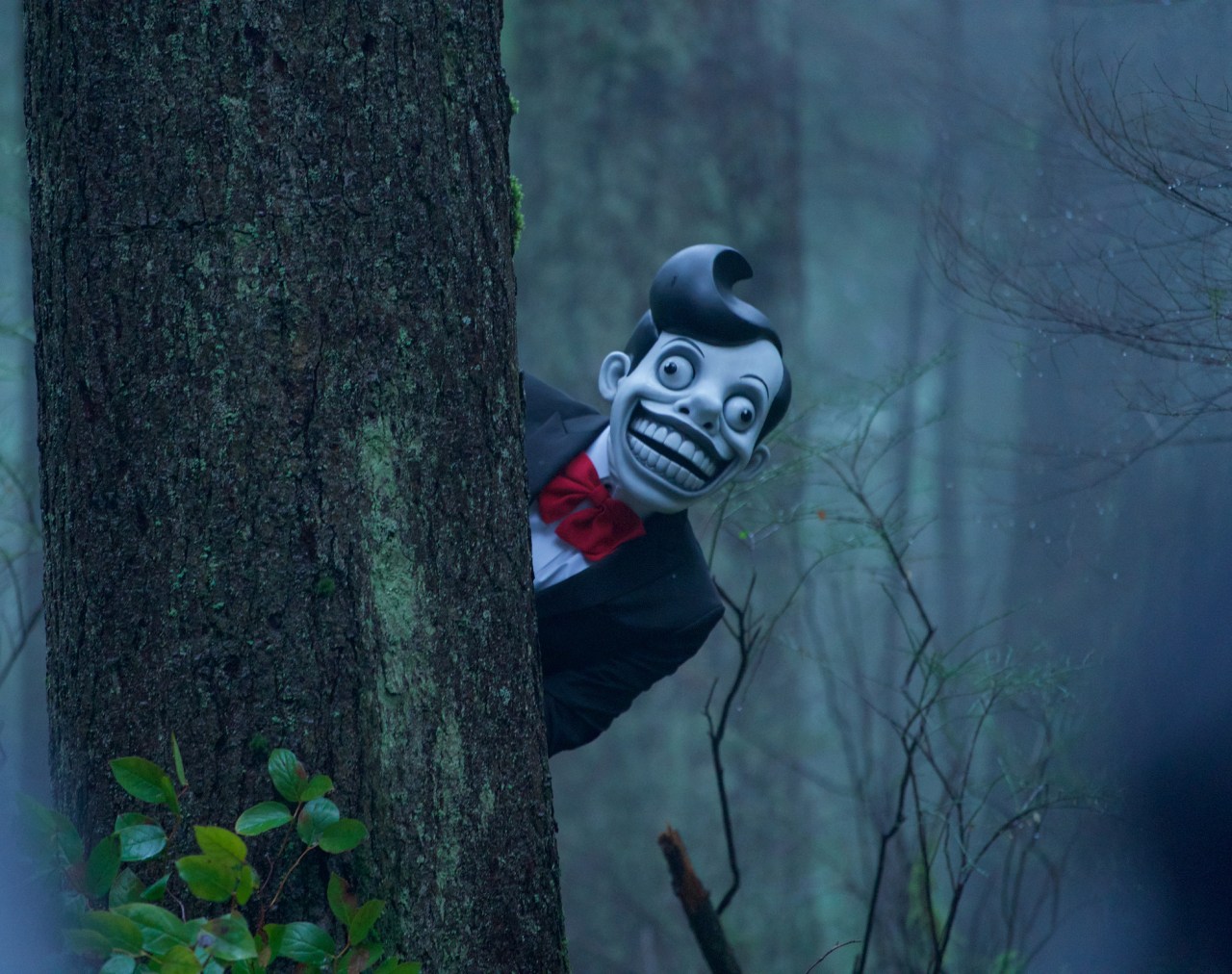 Creepy cartoon-like masked figure with a wide grin and red bow tie peeking around a mossy tree trunk in a dark, misty forest.