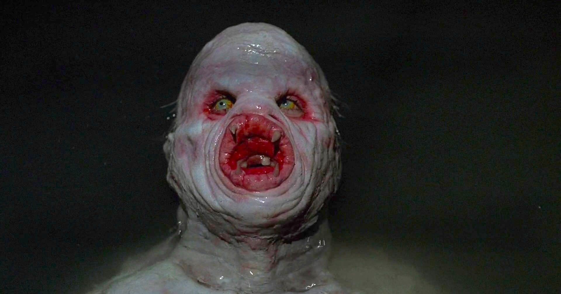 Close-up of a pale, grotesque humanoid creature with wrinkled skin, sunken yellow eyes, and a circular lamprey-like mouth full of teeth against a dark background.